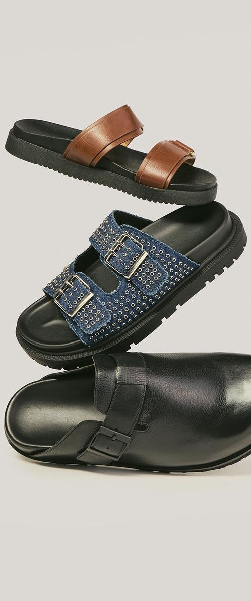 Drie slide sandalen: bruin leer met twee bandjes, blauw gestikt stof met gespen, en zwart leer met een zijgesp, verticaal gerangschikt.