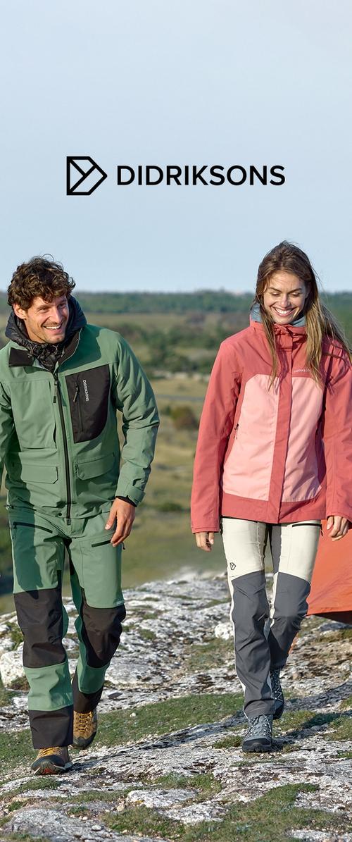 Dos personas caminan por un terreno rocoso, llevando una chaqueta impermeable verde con un bolsillo negro y otra chaqueta impermeable rosa, ambas con pantalones de contraste.