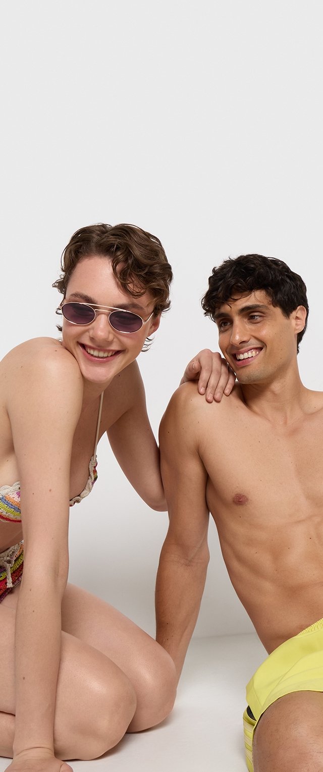 Dos modelos posan juntas en ropa de baño. Una lleva un bikini de rayas multicolores y gafas de sol moradas. La otra lleva unos pantalones cortos de baño amarillos.
