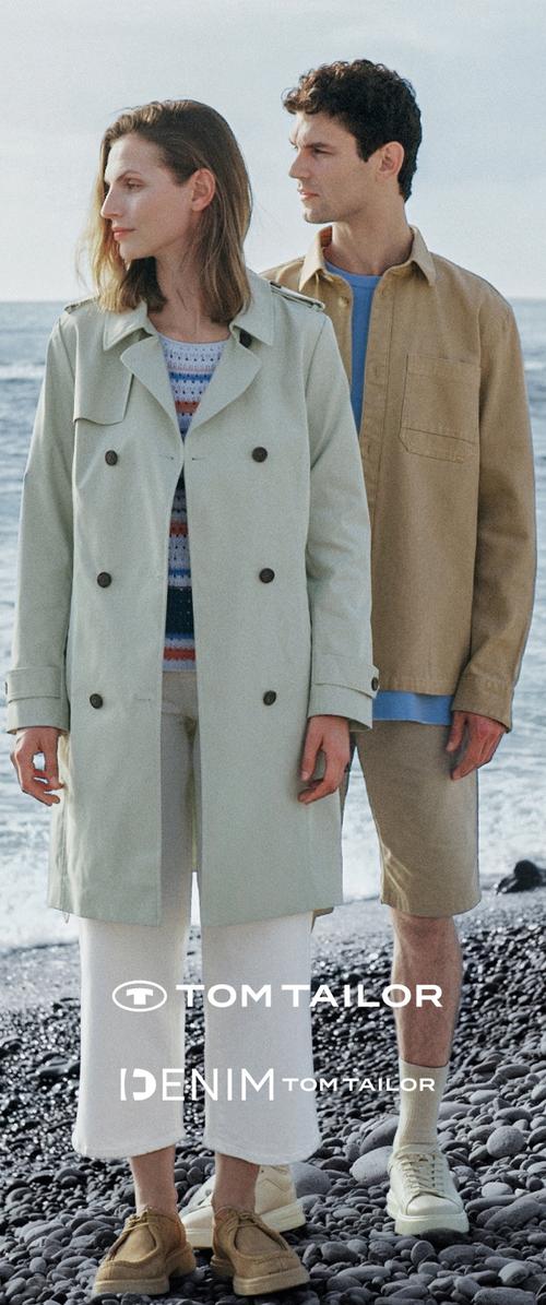 Hellgrüner Trenchcoat mit doppelreihiger Knopfleiste, kombiniert mit einem gestreiften Oberteil. Mann im beigen Hemd und Shorts; beide tragen neutrale Schuhe.