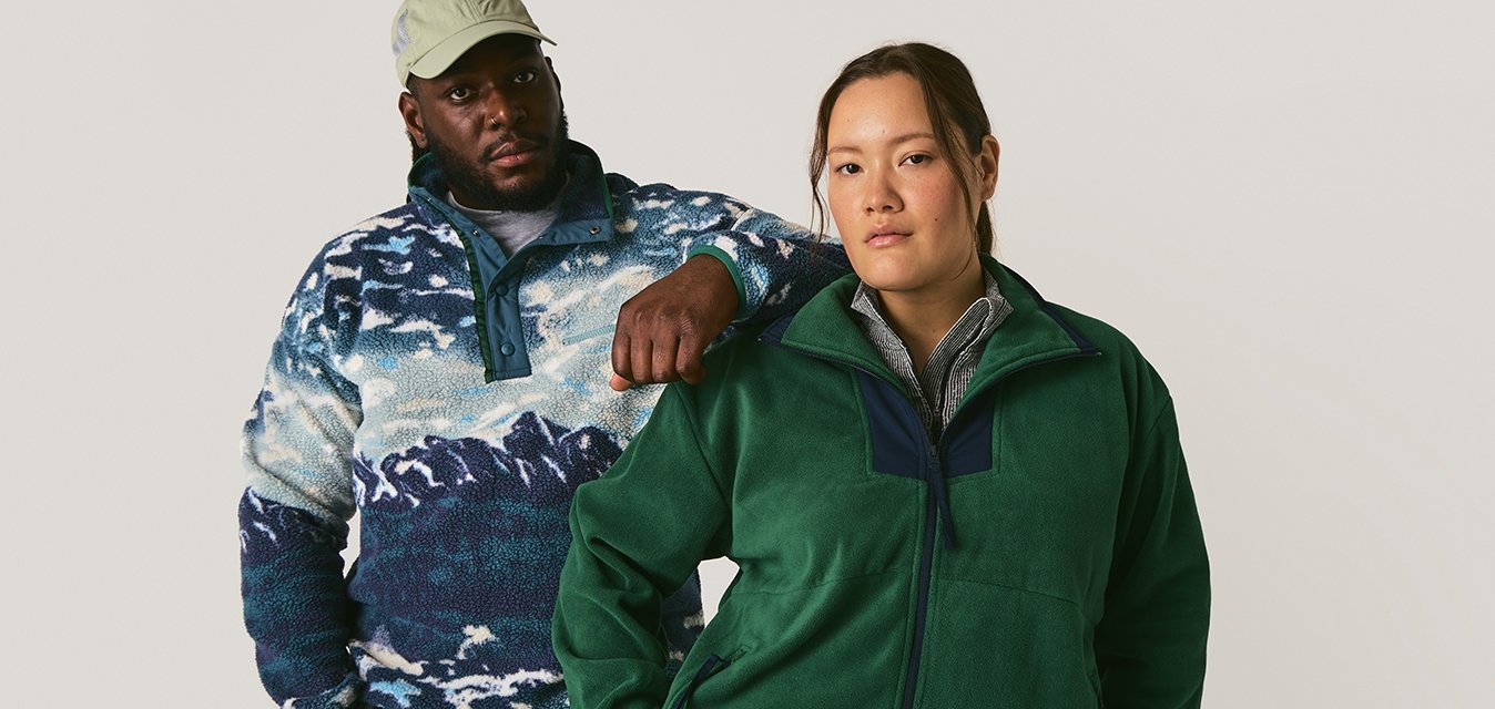 Muž v modrém vzorovaném fleecovém svetru a béžové kšiltovce stojí s rukou na rameni ženy v zeleném fleecovém svetru na zip a černých kalhotách.