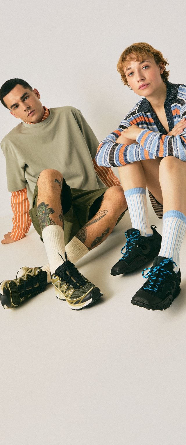 Deux jeunes adultes assis, portant des vêtements décontractés en couches et des baskets, avec des tatouages visibles et des chaussettes rayées sur un fond uni.
