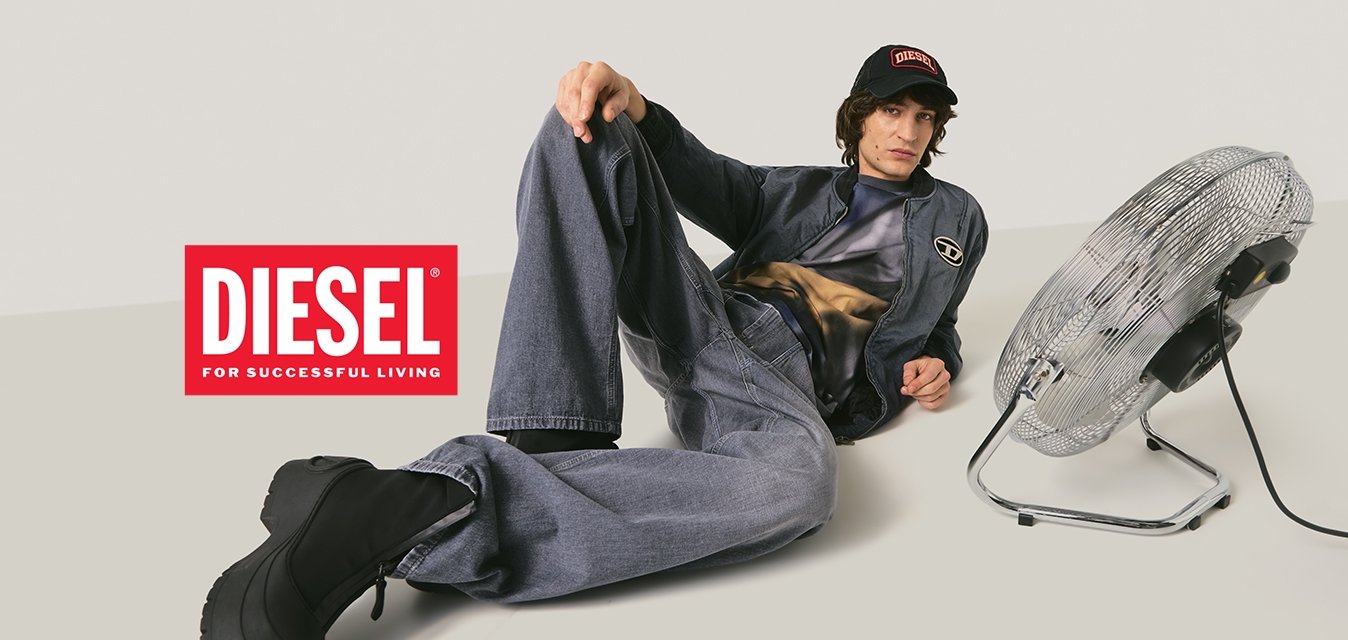 Osoba ve tmavém džínovém oblečení a černých botách leží na podlaze vedle kovového elektrického ventilátoru na jednobarevném pozadí.