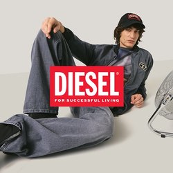 Osoba ve tmavém džínovém oblečení a černých botách leží na podlaze vedle kovového elektrického ventilátoru na jednobarevném pozadí.