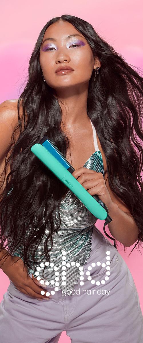 Frau mit langen, welligen Haaren hält ein türkisfarbenes Glätteisen, trägt ein glänzendes Paillettentop, vor rosa Hintergrund mit dem Text "ghd good hair day".
