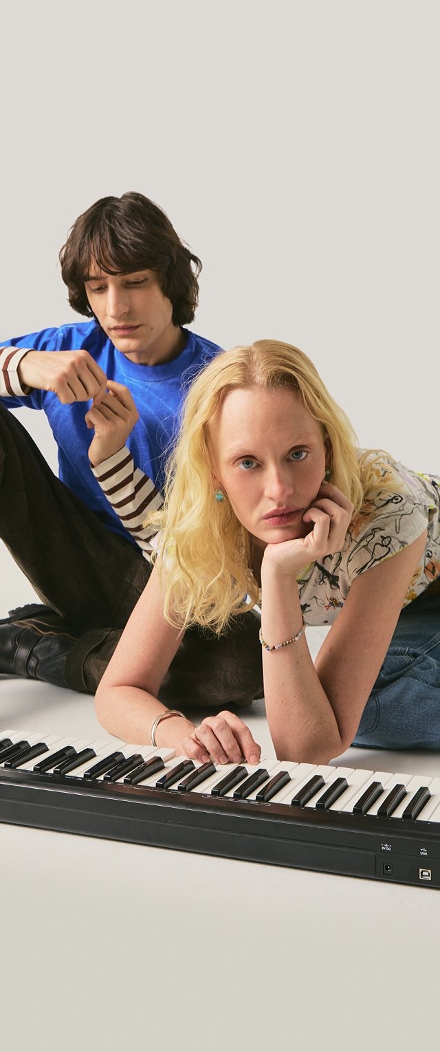 Joven mujer rubia tocando un teclado mientras un joven con camisa azul se sienta detrás de ella, ambos sobre un fondo claro y sencillo.