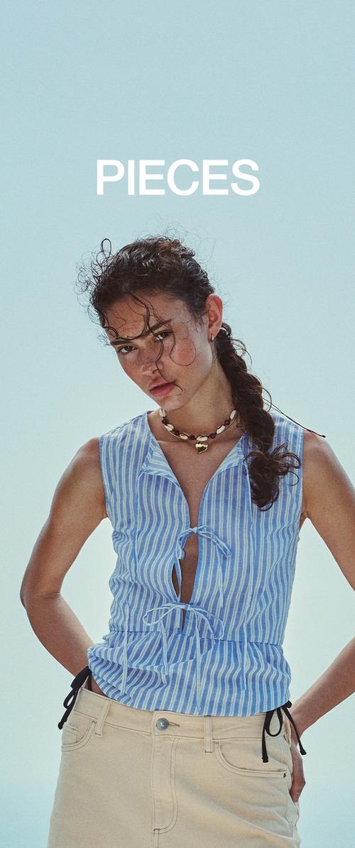Jonge vrouw met krullend haar in een gestreept blauw mouwloos topje en beige broek, draagt een schelpenketting, staand tegen een effen lichte achtergrond.