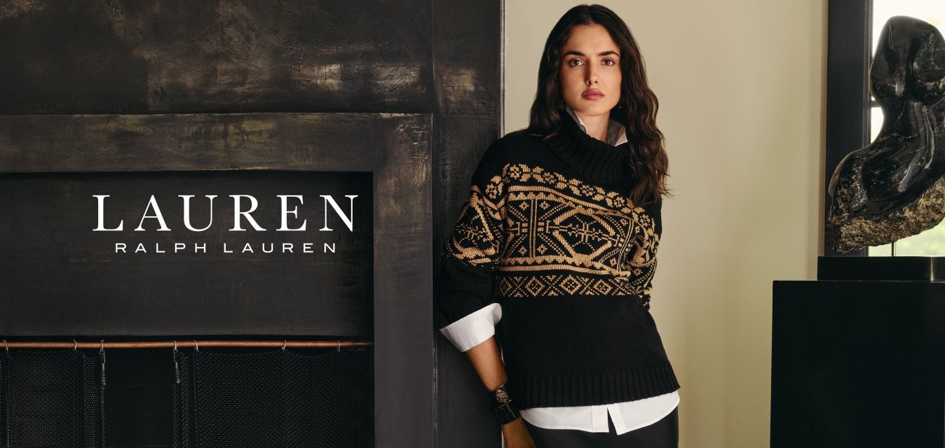 Žena v černém s béžovým vzorovaným svetrem a dlouhou černou sukní stojí u tmavého krbu a abstraktní sochy na podstavci u okna.