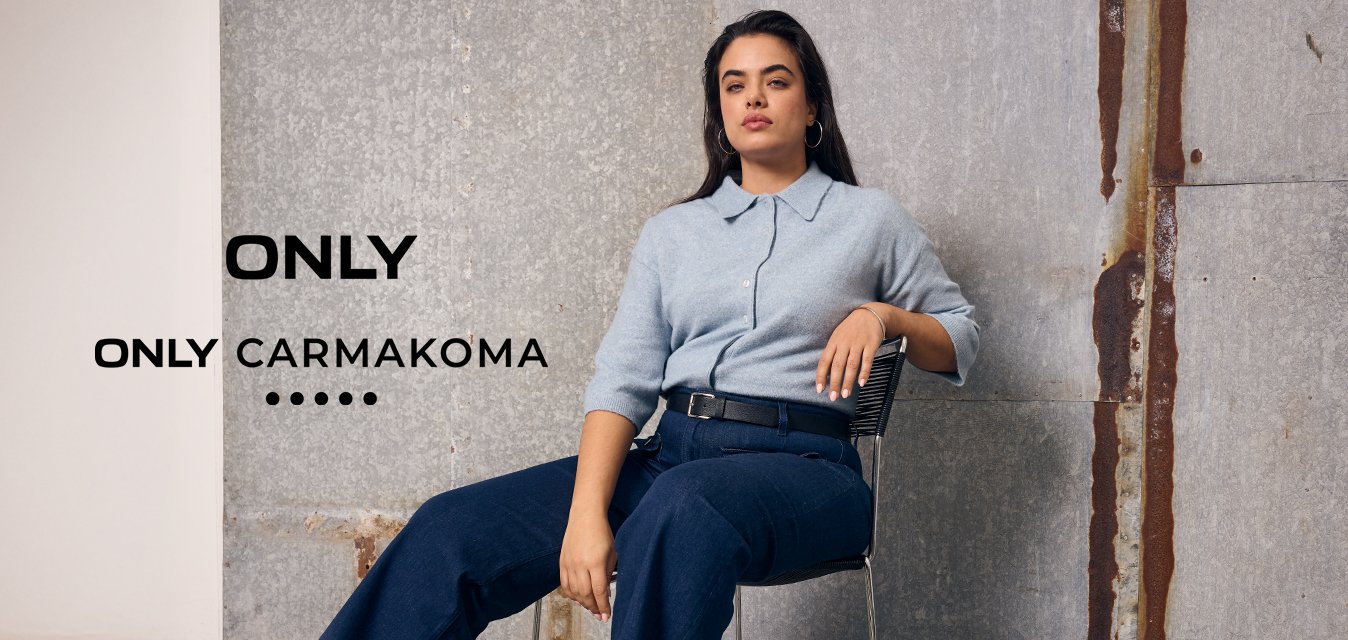 Žena s dlouhými vlasy sedí na židli, má na sobě světle modrý svetr, tmavě modré kalhoty a hnědé mokasíny, na pozadí šedé průmyslové zdi.