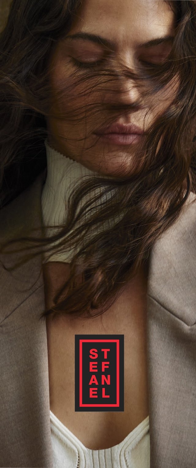 El cabello castaño cae sobre un suéter de cuello alto blanco y una chaqueta beige. Presenta un atrevido logo negro y rojo centrado en el pecho.