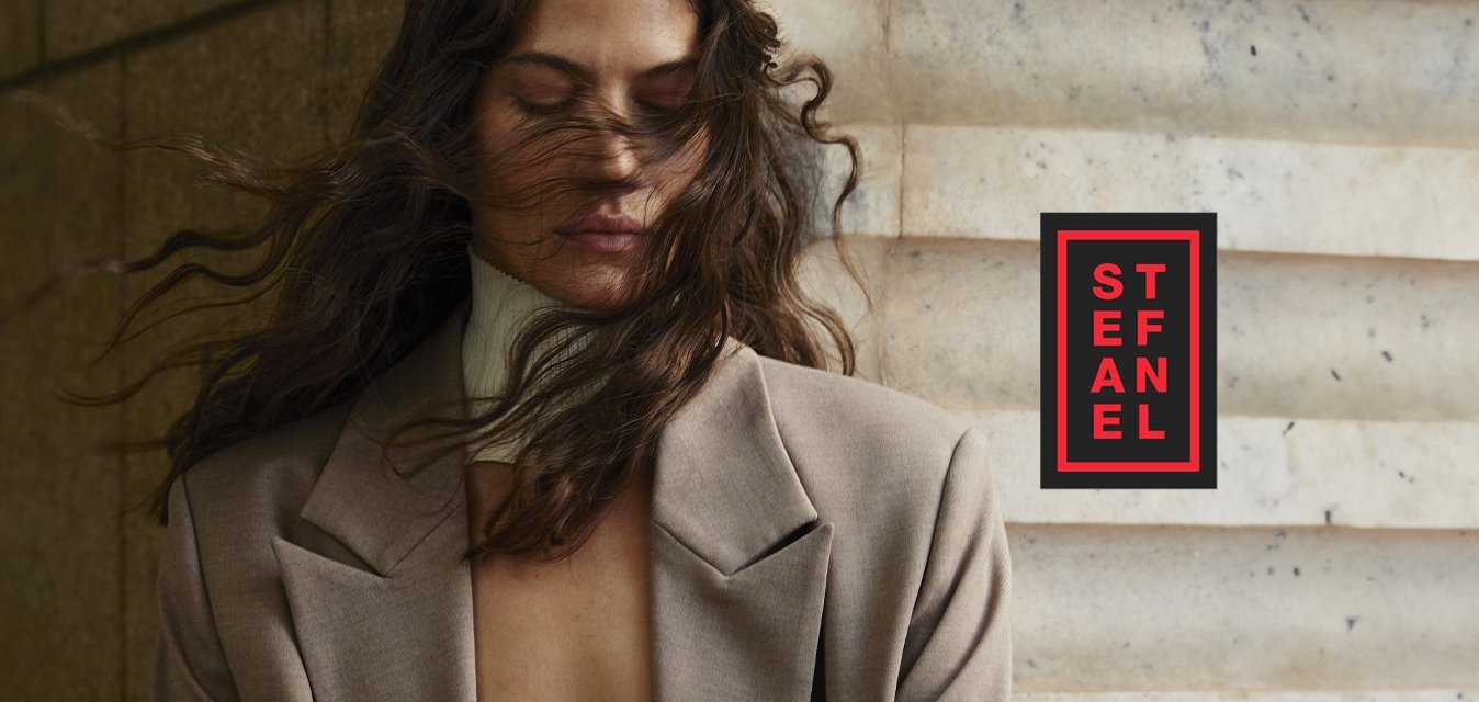 Bežový blazer s uvolněným střihem, jemnou texturou a ostrými klopami, v kombinaci s bílým žebrovaným topem. Vítr rozcuchané vlnité vlasy částečně zakrývají obličej.