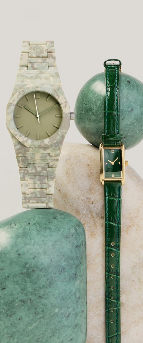 Deux montres vertes affichées verticalement sur des pierres lisses vertes et beiges, dont une avec un boîtier rectangulaire doré et un bracelet en cuir, et l'autre avec un bracelet camouflage.