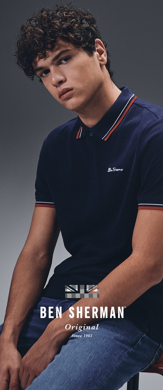 Námořnická modrá polo košile s žebrovaným límcem a červenými a bílými pruhy. Logo na hrudi; texturovaná látka; model sedící na šedém pozadí.