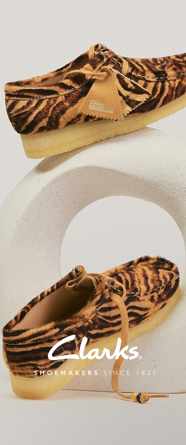 Paar Mokassins aus Wildleders mit Tigerfellmuster, mit Kreppsohlen, Lederbändern und einem hellbraunen Lederanhänger, präsentiert auf einem weißen, runden Ständer.