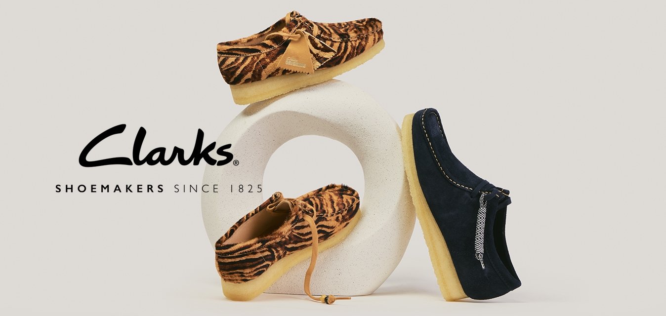 Dva hnědá mokasíny s tygřím vzorem a jeden tmavě modrý mokasín s béžovou podrážkou, vystavené na bílém sochařském podstavci.