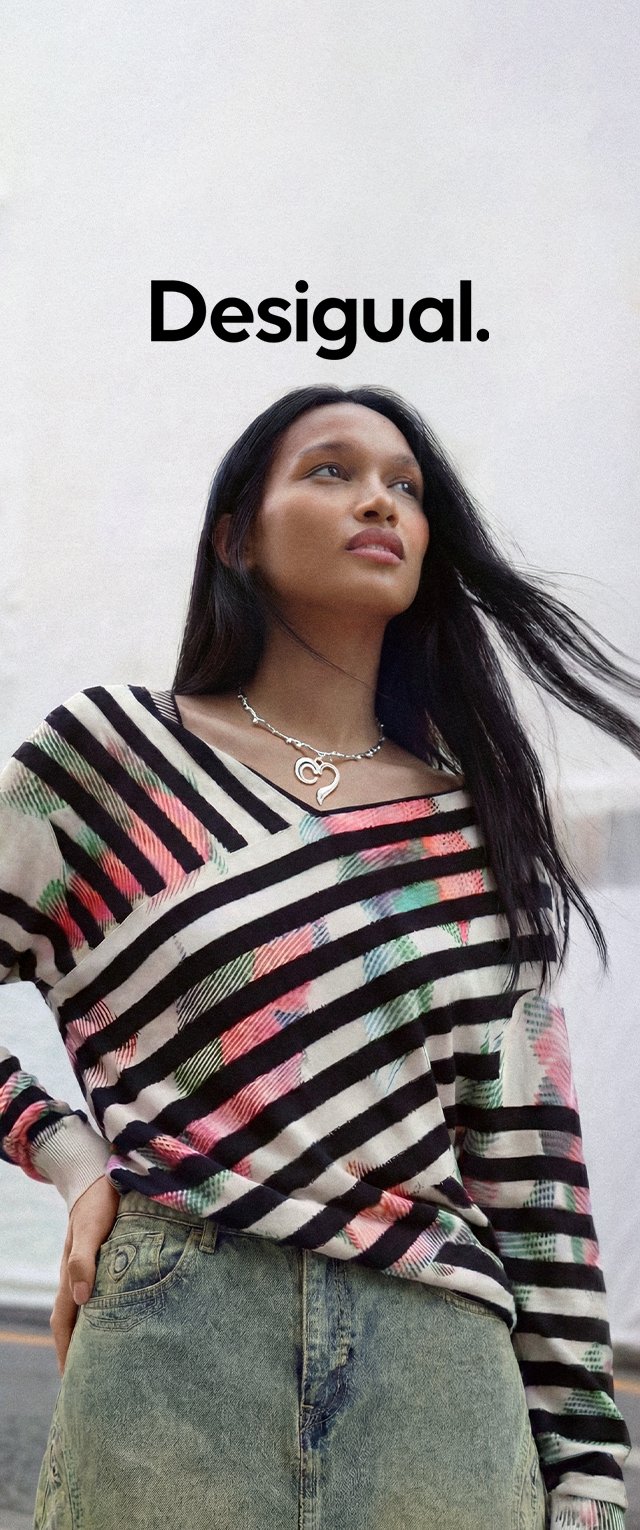 Joven mujer con cabello largo lleva un suéter a rayas y colorido y una falda verde de mezclilla, de pie al aire libre con un collar plateado en forma de corazón.