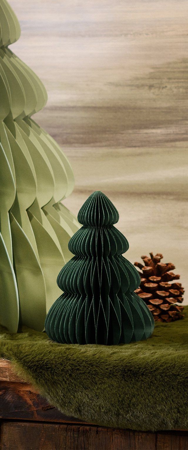 Grüner Papier-Weihnachtsbaum mit gefalteten Schichten, strukturierten Oberflächen und einer runden Basis, positioniert neben einem Tannenzapfen auf einer weichen grünen Felloberfläche.
