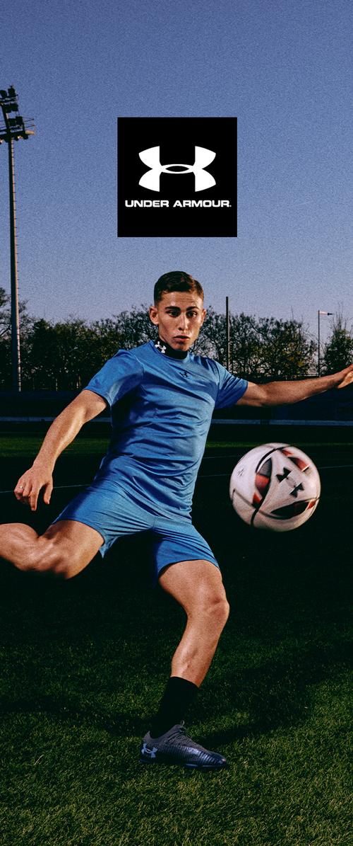 Jugador de fútbol masculino con ropa azul de Under Armour patea un balón de fútbol en un campo de césped bajo un cielo despejado al atardecer.