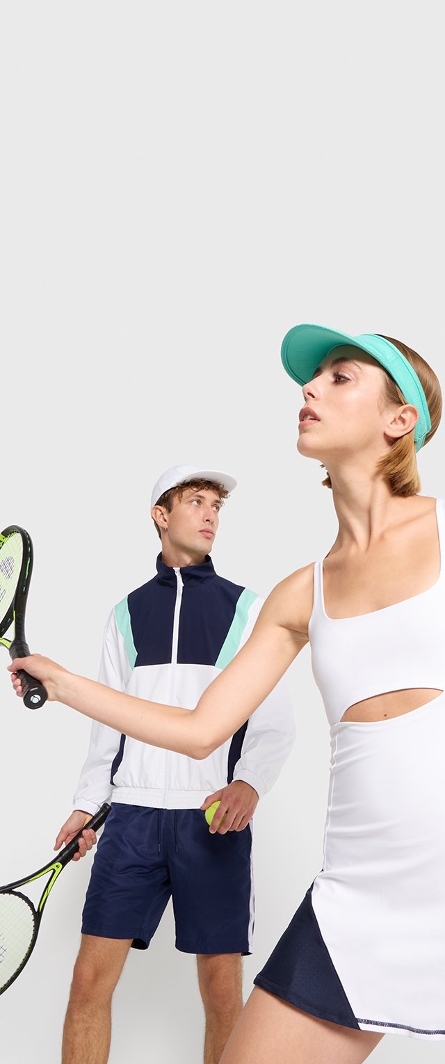 Canottiera sportiva bianca con dettagli a taglio, abbinata a pantaloni corti scuri. Uomo con giacca blu navy, cappello chiaro, che tiene una racchetta da tennis. Visiera verde come accessorio.