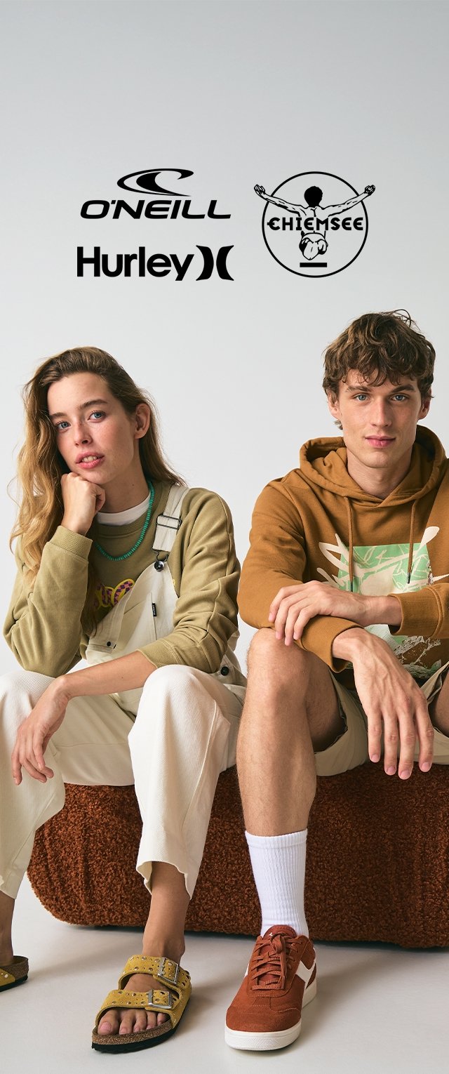Zwei Models sitzen auf einer strukturierten braunen Oberfläche. Die Frau trägt ein grünes Oberteil mit einem Logo, weiße Latzhosen und eine grüne Halskette. Der Mann trägt einen braunen Hoodie mit einem grafischen Design und orangefarbenen Sneakers.