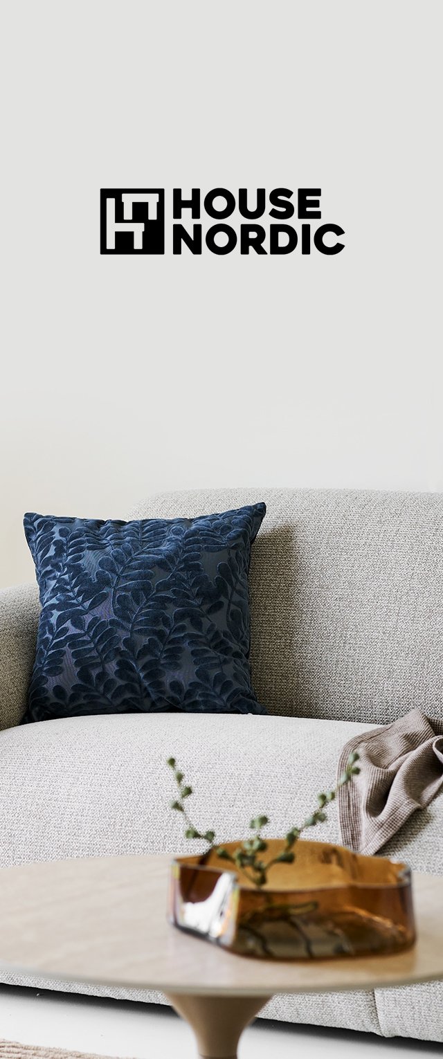 Sofá gris con textura, cojín azul oscuro con estampado de hojas, manta marrón, mesa de madera con jarrón de cristal ámbar que contiene tallos verdes. Logo de HOUSE NORDIC arriba.