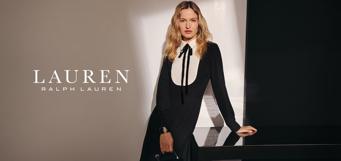 Žena s dlouhými blond vlasy má na sobě černé šaty s bílým límečkem ve tvaru bryndáčku, drží černou kabelku a stojí vedle tmavého lesklého stolu.