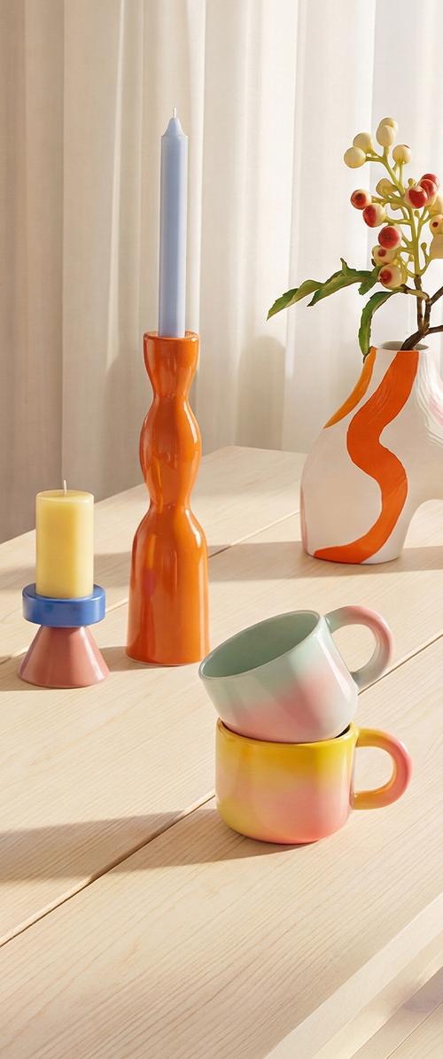 To gradienterede keramikkrus stablet, orange lysestage med blåt stearinlys, pink og blå lysestage med gult stearinlys, vase med bær på et træbord.