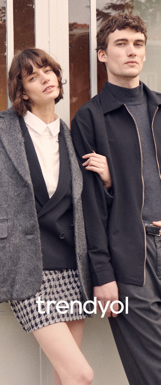 Cappotto grigio strutturato, giacca nera con zip e pantaloni corti a motivi. Camicia bianca sotto con colletto distintivo. Colori neutri, design moderno.