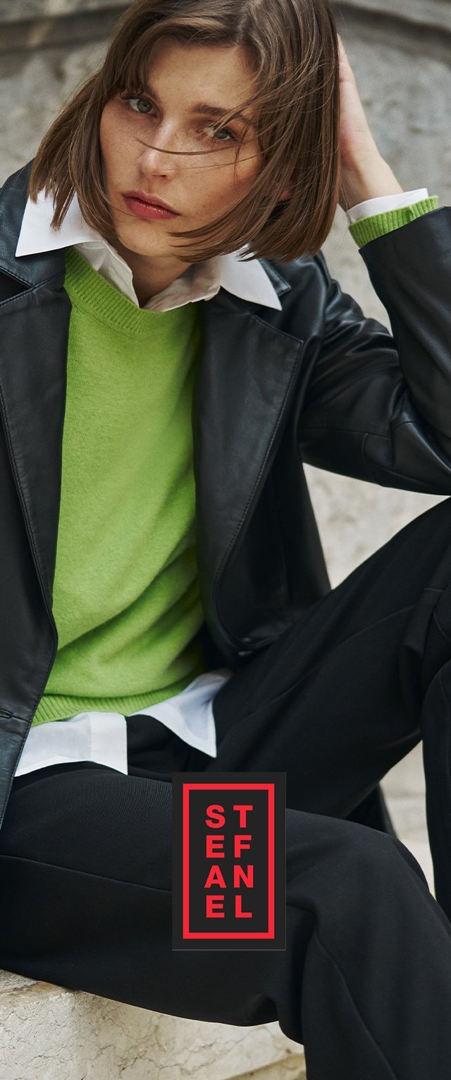 Zwarte leren jas over een felgroene trui met een witte kraag, gecombineerd met donkere broek. Logo in een rood en zwart rechthoek aan de onderkant.