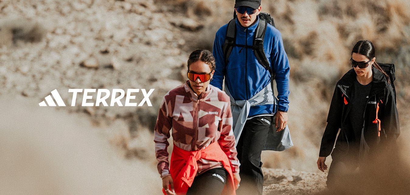 Tři turisté v outdoorovém oblečení a slunečních brýlích kráčejí po kamenité stezce v suché, travnaté krajině.