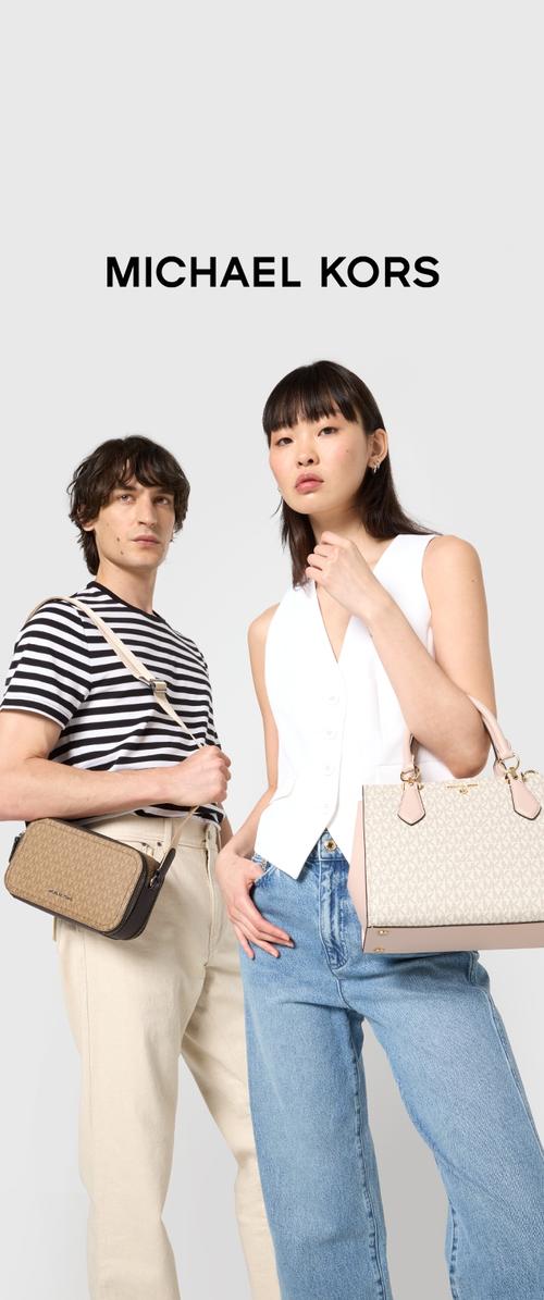 Dos modelos posan con bolsas de Michael Kors. La mujer sostiene un bolso de mano de patrón marrón; el hombre tiene un bolso cruzado marrón. Ambos visten ropa casual.