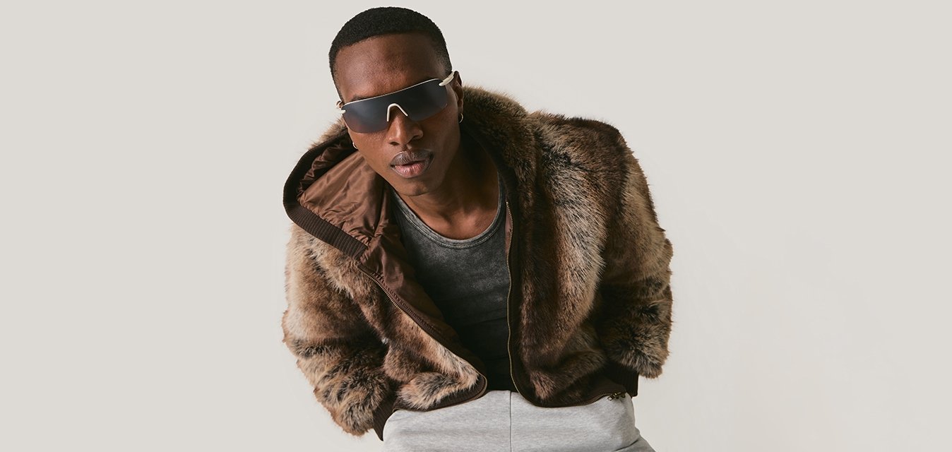 Mladý muž nosící tmavé sluneční brýle, hnědý kožich, šedé tričko a světle šedé kalhoty, nakloněný vpřed na jednoduchém pozadí.