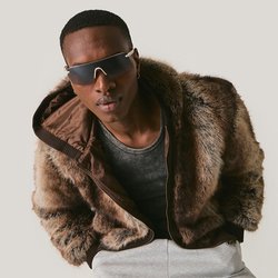 Mladý muž nosící tmavé sluneční brýle, hnědý kožich, šedé tričko a světle šedé kalhoty, nakloněný vpřed na jednoduchém pozadí.