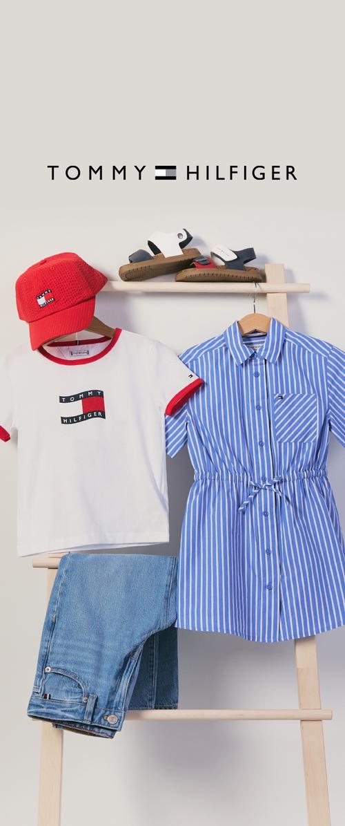 Punainen Tommy Hilfiger -lippis, valkoinen logo t-paita, sininen raidallinen mekko, farkut ja sandaalit esillä puisella telineellä, jonka yläpuolella on Tommy Hilfiger -logo.