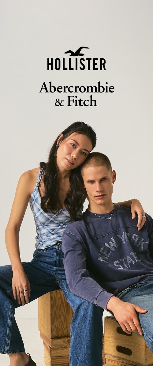 Joven mujer y hombre con atuendos casuales de mezclilla sentados sobre cajas de madera bajo los logotipos de Hollister y Abercrombie & Fitch sobre un fondo liso.