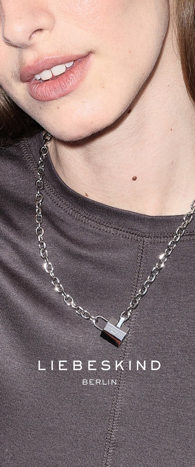 Silberne Metalldelnecklace mit einem Vorhängeschloss-Anhänger auf einem strukturierten grauen Kleidungsstück. Kettendesign mit Gliedern und einer glatten, polierten Oberfläche.