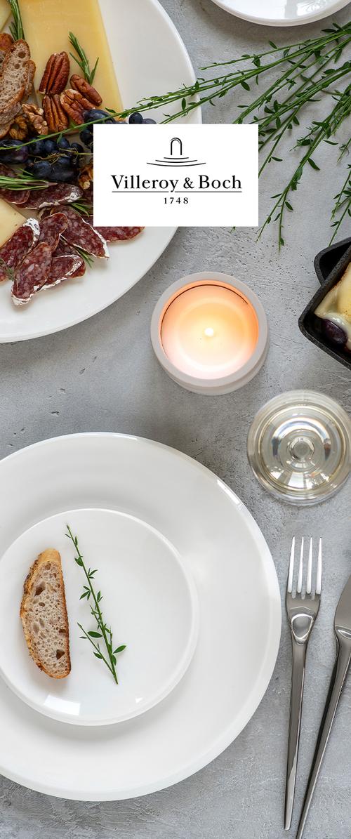 Charcuterie-lautanen, jossa viipaloitua salamia, juustoa, pähkinöitä, viinirypäleitä, leipää, yrttejä, valkoiset lautaset, haarukka, veitsi, kynttilä ja lasi harmaalla pöydällä.