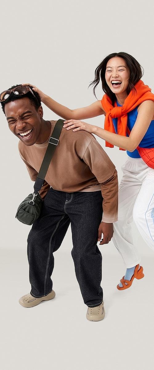 Dos jóvenes adultos riendo, uno inclinándose de forma juguetona hacia adelante con gafas de sol en la cabeza, el otro lleva una camiseta azul y un suéter naranja sobre los hombros.