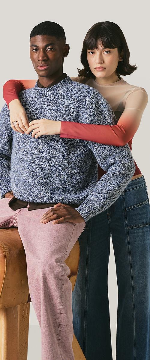 Un jeune homme portant un pull en tricot bleu et un pantalon rose est assis sur un banc en bois, une femme en haut rouge transparent et jean foncé se tient derrière lui, les bras autour de lui.
