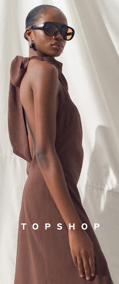 Woman wearing sleeveless brown halter dress with back tie and large dark sunglasses, standing against white fabric background.