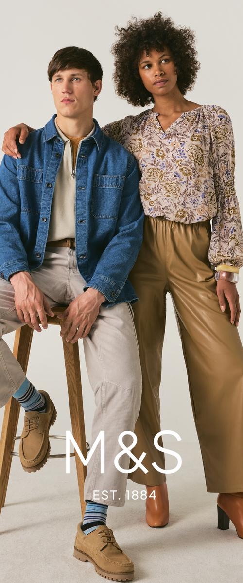 Hombre con chaqueta vaquera y pantalones beige sentado en un taburete; mujer de pie a su lado con blusa floral y pantalones de cuero marrón; fondo neutro.
