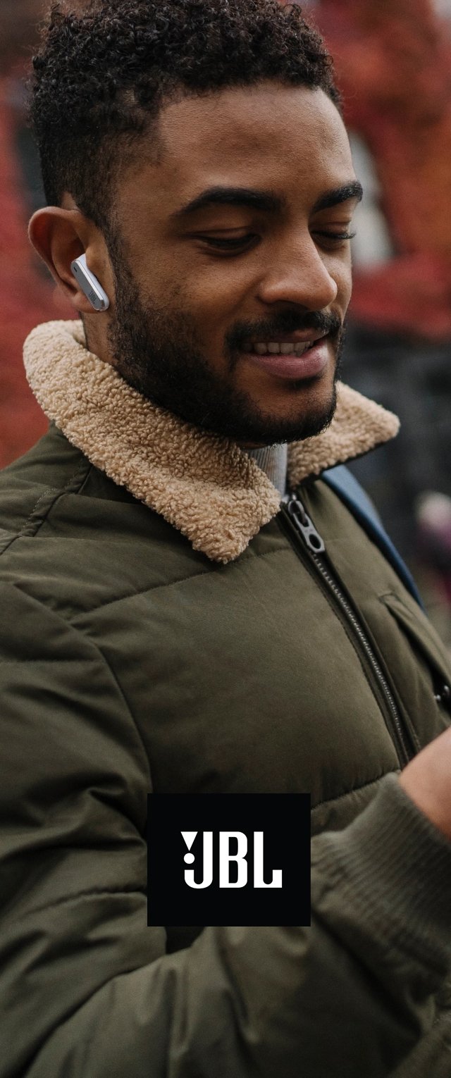 Mężczyzna noszący bezprzewodowe słuchawki JBL, uśmiechnięty i patrzący na ręczne urządzenie, ubrany w zieloną kurtkę z beżowym kołnierzem z polaru na świeżym powietrzu.