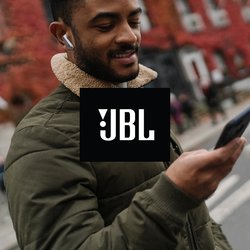 Muž v sluchátkách a zelené bundě hledí na chytrý telefon na městské ulici s budovami s červenými listy a zaparkovaným autem v pozadí.