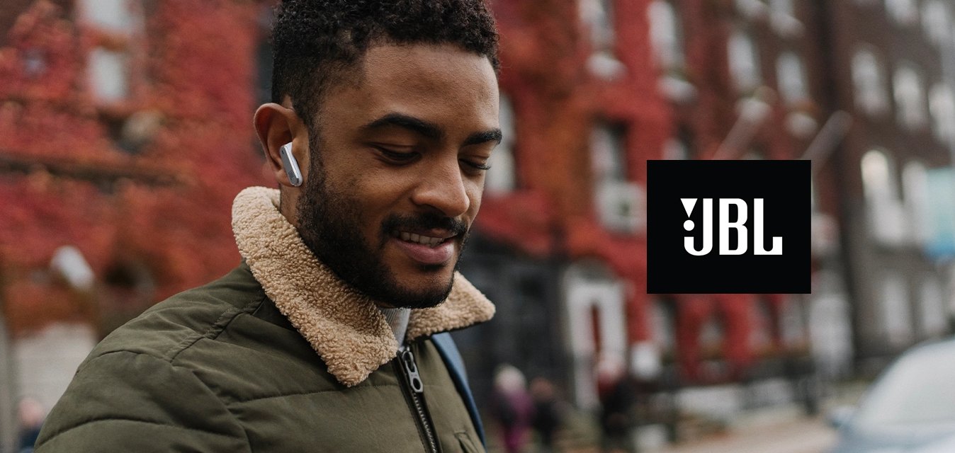 Muž v sluchátkách a zelené bundě hledí na chytrý telefon na městské ulici s budovami s červenými listy a zaparkovaným autem v pozadí.