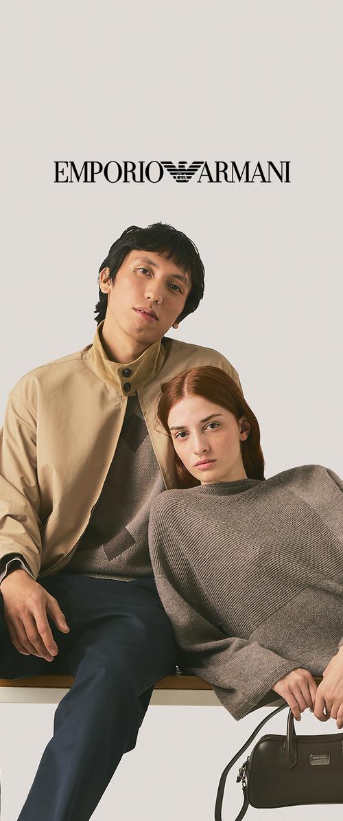 Hombre con chaqueta beige y mujer con suéter gris sentados juntos bajo el logo de Emporio Armani, con la mujer sosteniendo un bolso negro.