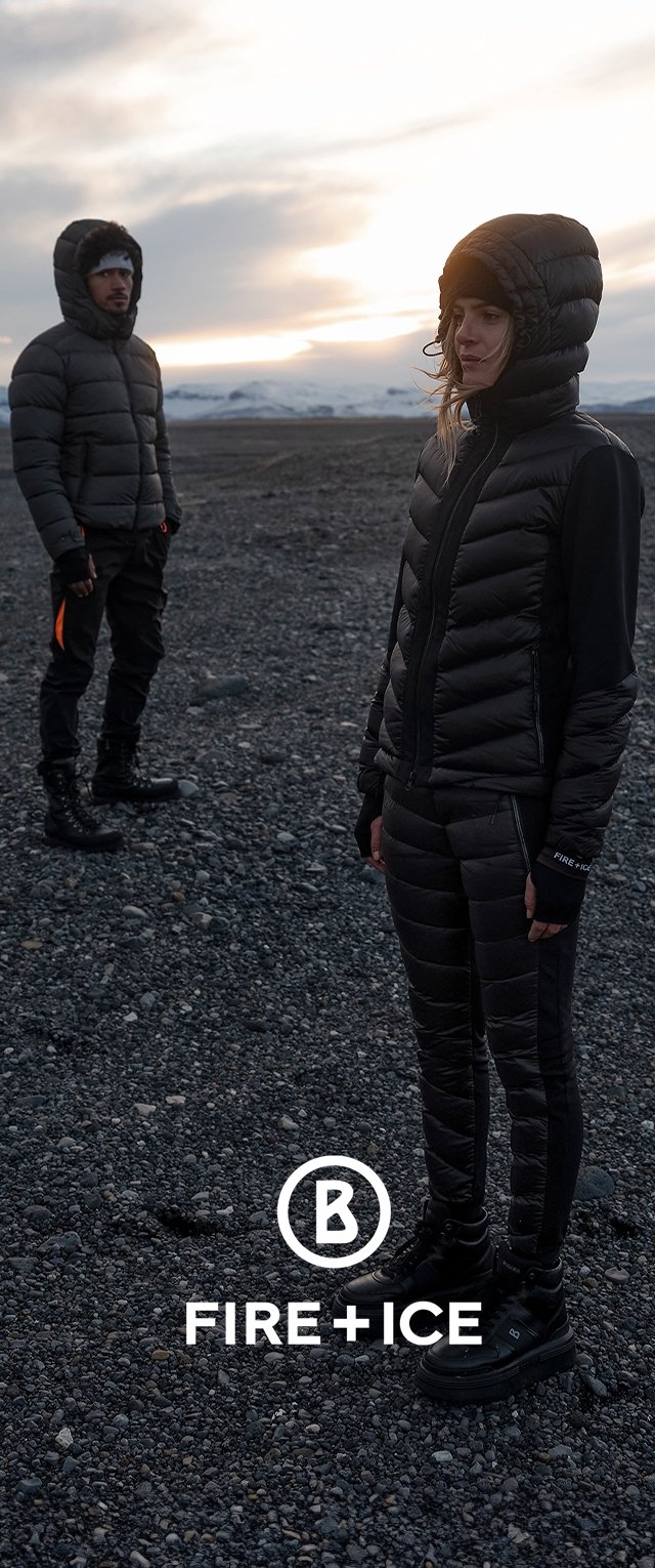 Divas figūras stāv uz akmeņainas virsmas, ģērbtas melnās izolētās jakās un biksēs, uz kurām apakšā redzams logo. Matēta tekstūra kontrastē ar ainavu.