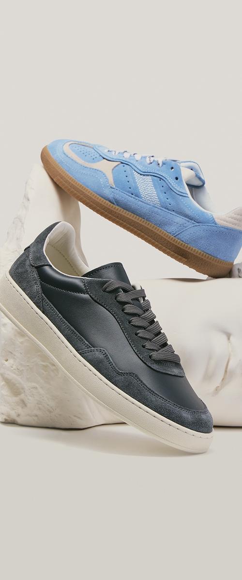 Twee casual sneakers, één lichtblauw met een gumzool en één zwart met een witte zool, tentoongesteld op witte gebeeldhouwde standaards tegen een neutrale achtergrond.
