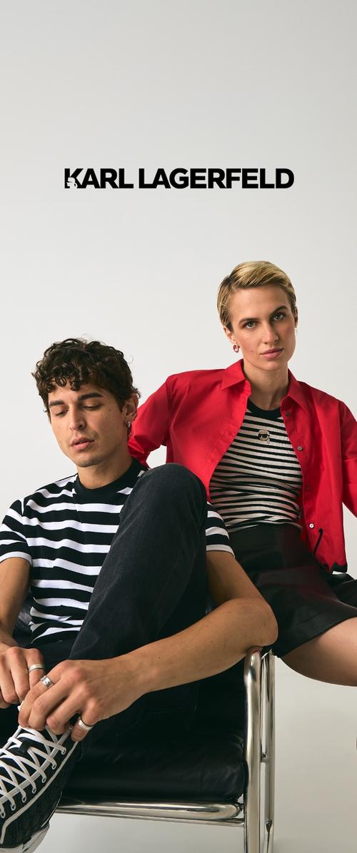 Model wearing a red shirt over a black and white striped top, seated on a black chair. Backdrop is plain white, enhancing the clothing contrast.