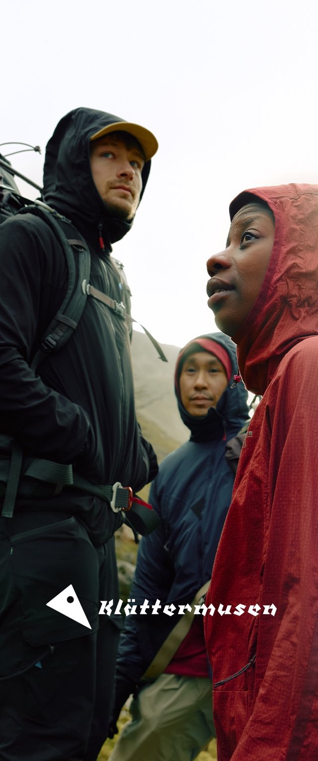 Tres personas con ropa de exterior: una con chaqueta gris y negra, otra con chaquetas roja y negra, y mochilas, en un fondo brumoso.