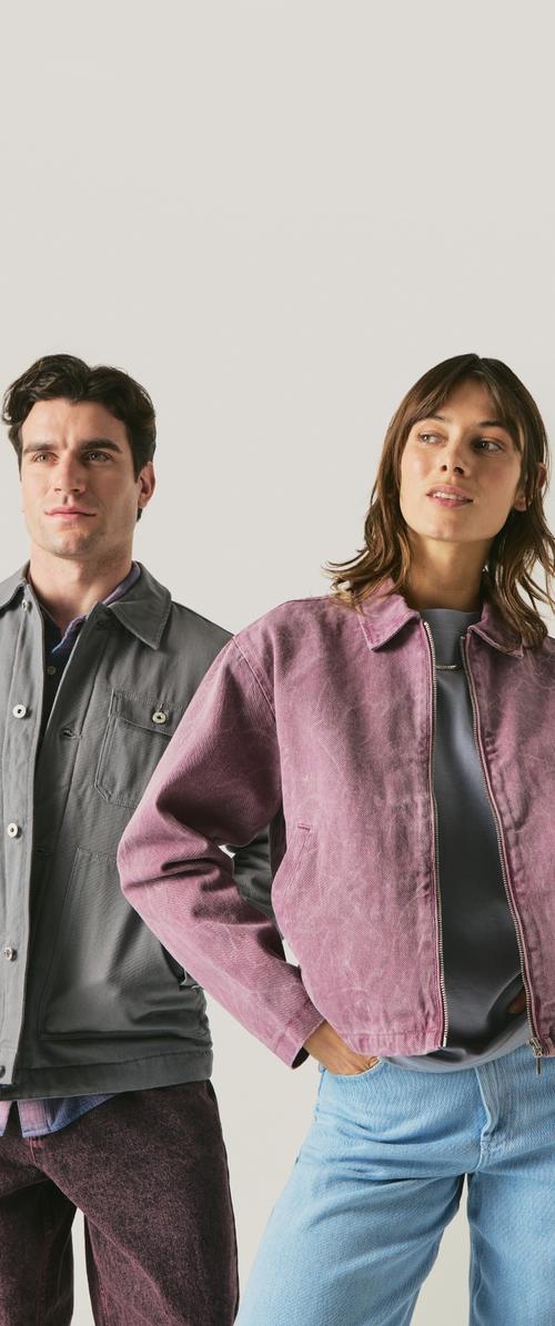 Hombre con chaqueta gris y pantalones oscuros de pie junto a mujer con chaqueta rosa y vaqueros azul claro sobre un fondo liso y claro.